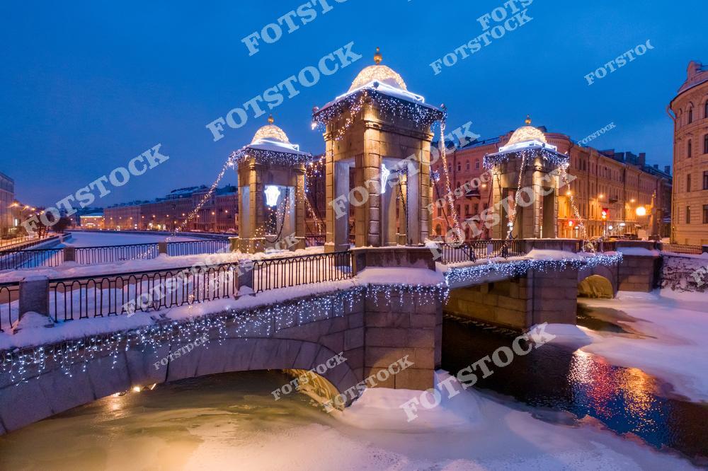 Фотография зимнего ночного Санкт-Петербург, Россия, сделанная в период рождественских и новогодних каникул. На изображении запечатлён Ломоносовский мост, украшенный праздничной иллюминацией и новогодним декором. Фотоснимок передаёт атмосферу зимнего путешествия по России: снег, огни ночного города и отражения света в воде создают ощущение уюта и праздника. Фото подчёркивает привлекательность рождественских туров в Санкт-Петербург и красоту города в зимний сезон. Иллюстрация может использоваться как изображение, картинка или слайд, посвящённые новогодним путешествиям и зимнему Петербургу.